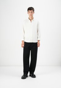 Camisa abotonada blanca texturizada de manga larga, combinada con pantalones negros holgados y zapatos negros de suela gruesa.
