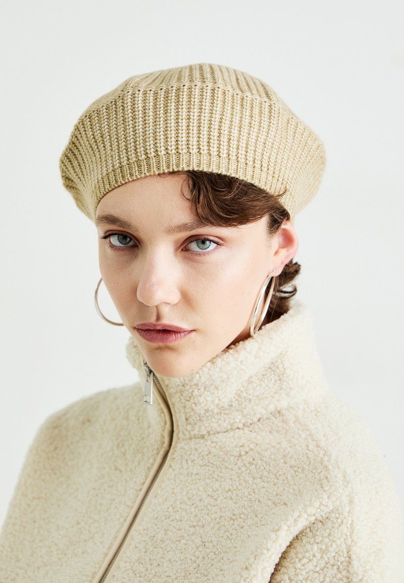 Beige Rippstrick-Beret mit weicher Textur. Getragen mit einer cremefarbenen, fuzzy Reißverschlussjacke, die einen hohen Kragen und einen glatten Reißverschluss hat.