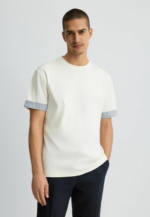 Kurzärmliges, cremefarbenes T-Shirt mit gerippten grau-blauen Bündchen. Weicher Stoff, lockere Passform, Rundhalsausschnitt. Getragen mit dunkelblauen Hosen.