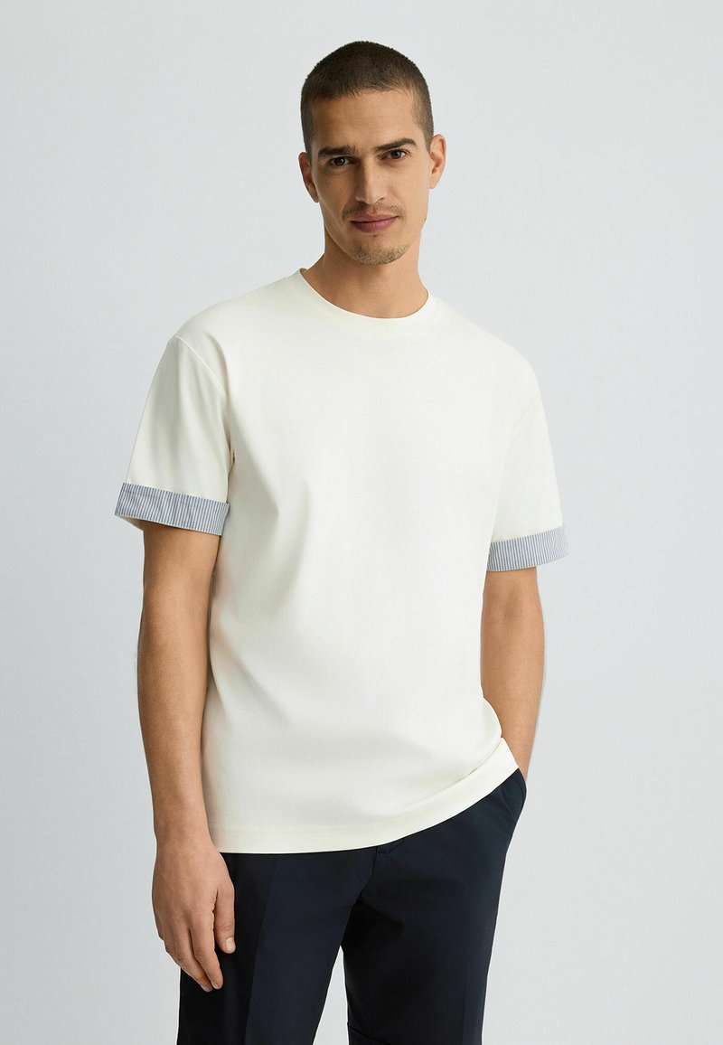 Camiseta de manga corta color crema con puños en gris azulado acanalados. Tejido suave, corte holgado, cuello redondo. Llevada con pantalones azul marino oscuros.