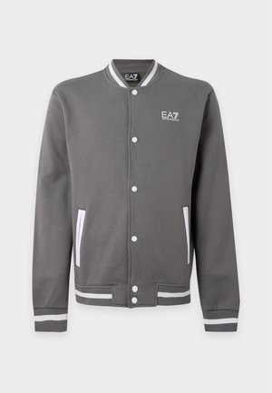 Jeune homme portant une casquette beige, une veste universitaire grise, un t-shirt vert, un pantalon beige et des baskets blanches, debout devant un fond uni.