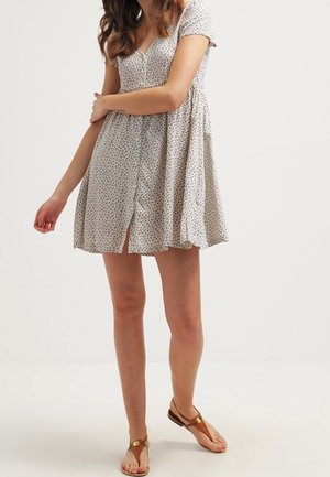 Frau trägt ein kurzärmliges, geblümtes Kleid mit Knopfleiste und V-Ausschnitt sowie braune Zehensandalen und steht vor einem einfarbigen Hintergrund.