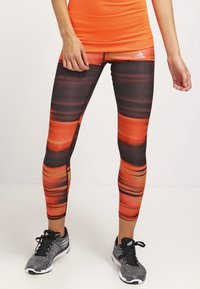 Leggings Adidas avec un motif de rayures horizontales orange vif et noir, fabriquées en tissu extensible pour le confort, associés à des chaussures de sport grises.