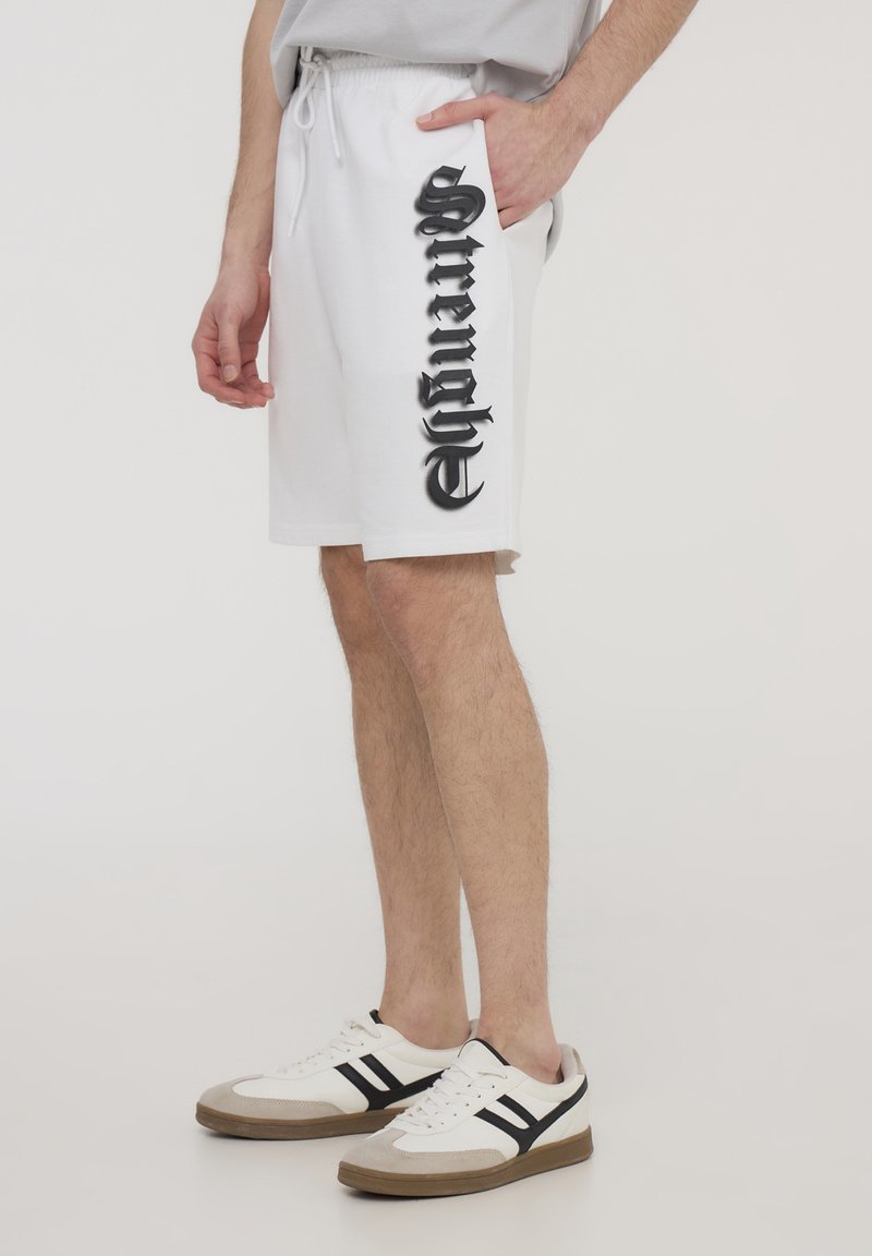 Shorts bianchi in cotone con un design grafico nero laterale, dotati di vita elastica e tasche laterali. Abbinati a sneakers bianche.