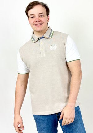 Jeune homme souriant portant un polo beige avec des manches blanches et un jean bleu, debout devant un fond blanc uni.