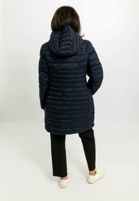 Cappotto imbottito blu navy con cappuccio, caratterizzato da un design trapuntato e una lunghezza media, indossato sopra pantaloni neri e scarpe chiare.