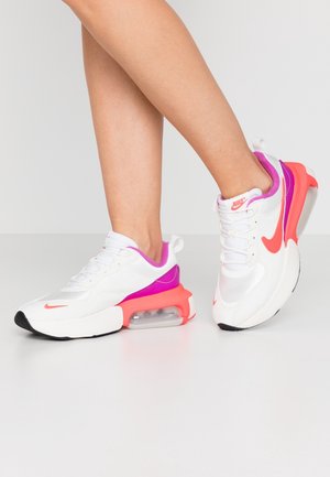 Jambes portant des baskets blanches Nike avec des touches rose et orange, un pied à plat et l'autre levé sur le talon, sur un fond uni.