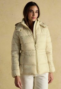 Krémová prešívaná bunda s predným zipsom, elastickými manžetami a patentkami na pleciach. Hladká textúra a horizontálny prešívaný dizajn.