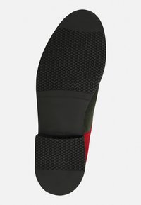 Schuhsohle mit einer strukturierten, schwarzen Gummisohle und einer glatten Ferse; rote und olivfarbene Wildlederakzente an den Seiten.