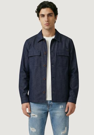 Mies pukeutuneena tummansiniseen napitettuun takkiin valkoisen paidan päällä, yhdistettynä vaaleansinisiin kuluneisiin farkkuihin, seisoo yksivärisen taustan edessä.