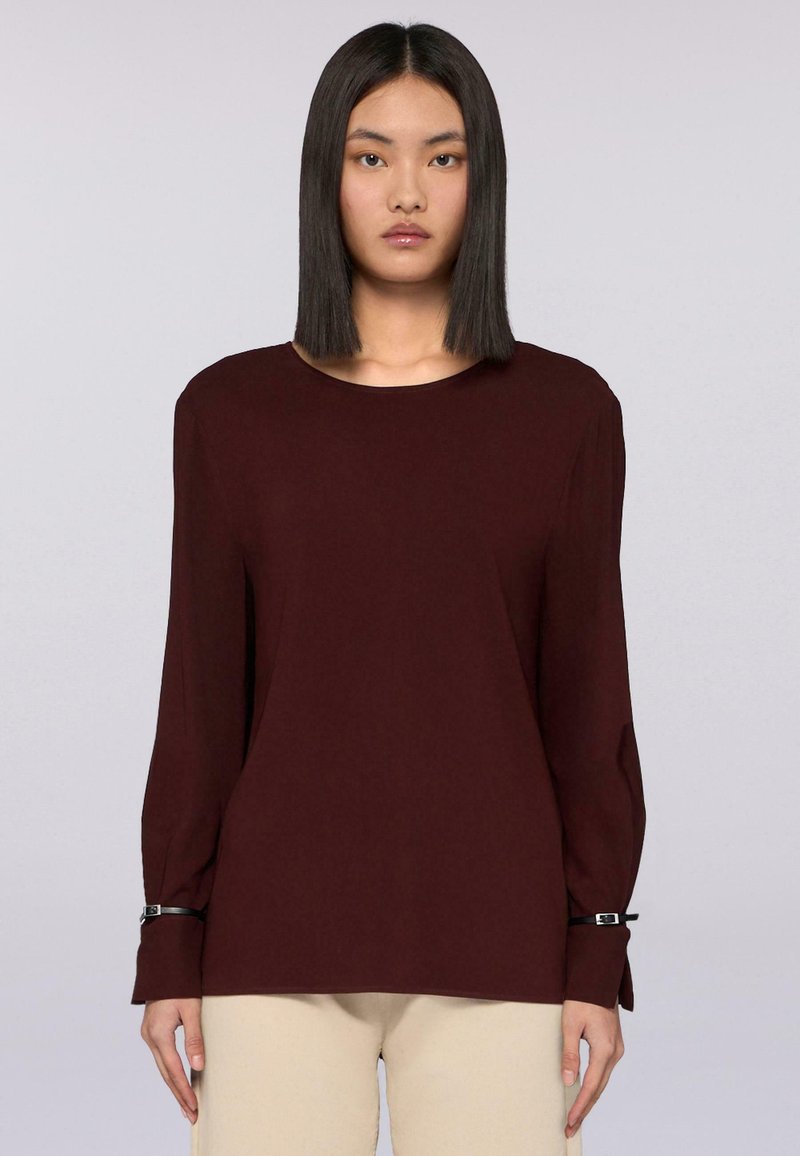 Donna che indossa una blusa a maniche lunghe di un rosso borgogna scuro con dettagli neri alle maniche e pantaloni beige, in piedi su uno sfondo semplice.