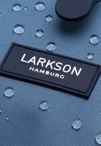 Modrá textúrovaná tkanina s vodnými kvapkami, obsahujúca čiernu nepriehľadnú etiketu s embosovaným nápisom "LARKSON HAMBURG" v bielych písmenách.