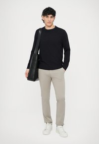 Juodas megztas megztinis, smėlio spalvos siauri kelnės, balti sportbačiai ir juoda peties rankinė. Stovi viena, žiūrėdama į kamerą priešais paprastą foną.