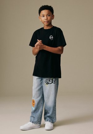 Băiat stând cu mâinile împreunate, purtând un tricou negru cu un design de craniu și blugi albastre deschis cu pete grafice colorate și adidași albi.