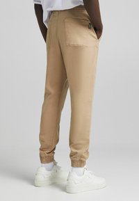Beige Baumwoll-Jogginghosen mit elastischen Bündchen und einer Gesäßtasche, kombiniert mit weißen Sneakers und sichtbaren weißen Socken.