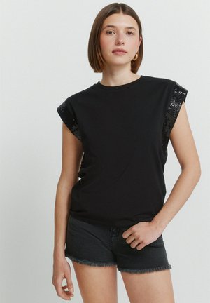 Nuori nainen, jolla on lyhyet ruskeat hiukset, hänellä on musta hihaton toppi, jossa on paljettihihat, ja mustat farkkushortsit yksiväristä taustaa vasten.