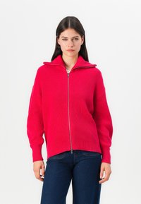Fuchsia ritssluiting sweater met ribbelstructuur, hoge kraag en een losse pasvorm. Gecombineerd met donkerblauwe denim jeans.
