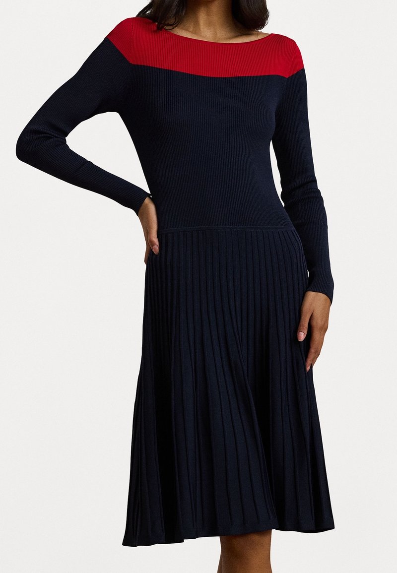 Femme portant une robe en maille côtelée bleu marine avec un large col rouge et des manches longues, jupe plissée sous la taille.