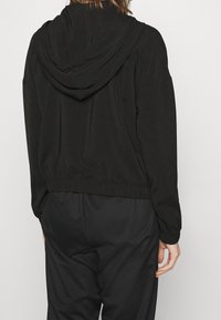 Svart kort hoodie gjord av mjukt material med dragsnodd i huvan och elastisk midja, sedd bakifrån. Enkel, minimalistisk design.