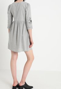 Grå, knälång klänning med trekvartslånga ärmar, som har blommönsterbroderi på ärmarna, rynkad midja och subtilt texturerad tyg.