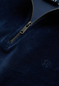 Sudadera con cremallera en color navy, de tejido suave; cuenta con una cremallera metálica y un pequeño logo bordado en el pecho. Textura suave, diseño casual.