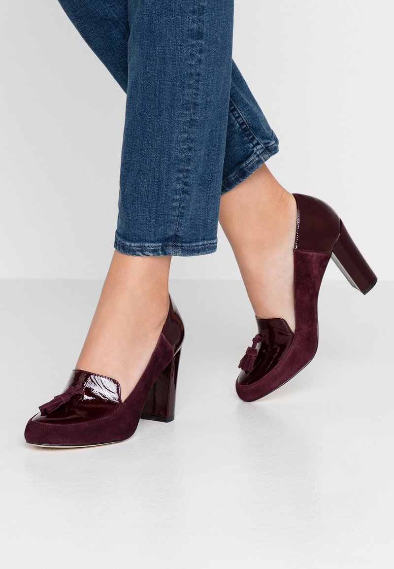 Femme portant un jean bleu et des mocassins à talon bloc en daim et cuir verni bordeaux avec détail à pampilles, debout sur un sol clair.