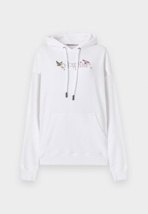 Frau trägt übergroßen weißen Hoodie mit Vogelmotiven, schwarze Cargohose mit Reißverschlüssen und Schnallen, schwarze Schuhe und große Sonnenbrille, stehend.