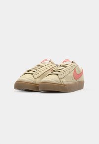 Beige suede sneakers with pink Swoosh logo, lace-up design, flat gum sole, and contrasting white tongue tag. Textured upper surface.