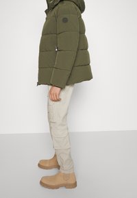 Prošívaná bunda v tmavě zelené barvě s kapucí a prošívaným designem. Kombinováno s lehkými béžovými cargo kalhotami a opálenými kotníkovými botami s texturovanou podešví.