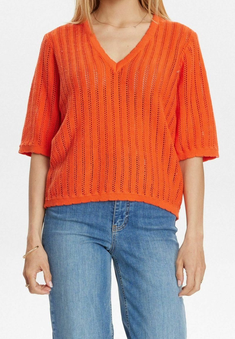 Femme portant un pull en maille orange vif à manches courtes et col en V, et un jean bleu, avec peu de bijoux aux mains et au cou.