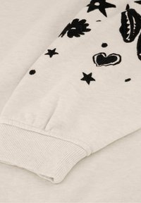 Baumwollpulloverärmeln in Off-White, mit schwarzen gestickten Blumen- und Sternmustern sowie einem gerippten Bündchen für zusätzliche Textur.