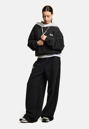 Młoda kobieta regulująca sznurek czarnej, oversize'owej bluzy z szerokimi czarnymi spodniami i sportowymi butami na jednolitym tle.