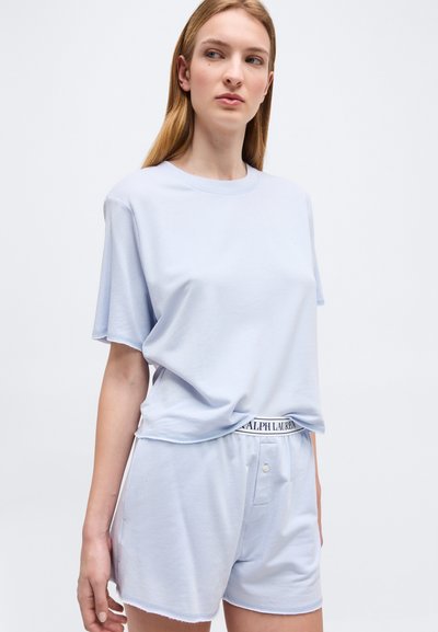 Γυναίκα που φοράει ανοιχτό μπλε κοντομάνικο πουκάμισο και κατάλληλο σορτς με ορατή ζώνη Ralph Lauren, στεκόμενη μπροστά σε απλό φόντο.