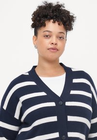 Τηλεγένη και λευκή ριγέ ζακέτα με V λαιμόκοψη, τρία κουμπιά και μαλακή, πλεκτή υφή. Χαλαρή εφαρμογή με φαρδιές μανσέτες.