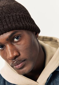 Berretto marrone lavorato a maglia con una texture a coste indossato da un modello. Il modello indossa una felpa con cappuccio chiara e una giacca di jeans, con un primo piano sui tratti del viso.