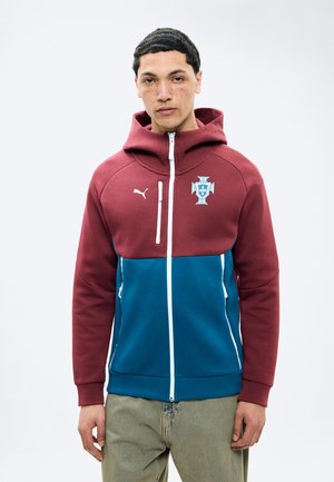 Νέος άνδρας που φοράει μια ζακέτα Puma με φερμουάρ σε μπορντό και μπλε χρώμα, με το έμβλημα της πορτογαλικής ποδοσφαιρικής ομάδας στο στήθος, στέκεται μπροστά σε μια απλή φόντο.