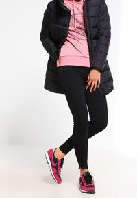 Persona que lleva una chaqueta acolchada negra, una sudadera con gráfico de búho rosa, pantalones skinny negros y zapatillas deportivas rosas y negras, de pie con las piernas cruzadas.