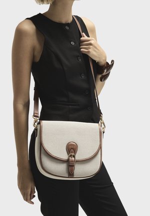 Bežová plátěná křížová taška s hnědými koženými detaily, zakřiveným klopou, přezkou a nastavitelným popruhem, kterou drží osoba ve černém topu.