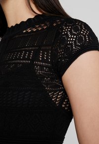 Černý pletený top s texturovaným krajkovým designem a geometrickými vzory, s vlnkovým výstřihem a krátkými rukávy.