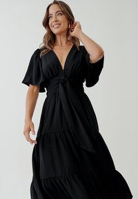 Black maxi dress with short sleeves, deep V-neckline, knotted waist detail, and tiered skirt. Made from smooth fabric with a flowing design.