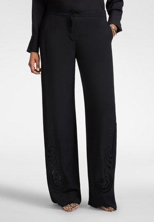 Kobieta ubrana w czarne szerokie spodnie z dekoracyjnymi wycięciami przy kostkach oraz błyszczące buty na spiczastym nosku.