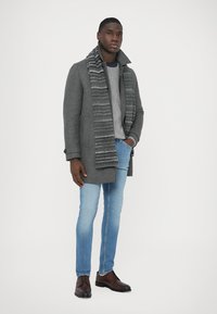 Palton gri din lână, fular gri cu model, pulover gri deschis, jeans albaștri și pantofi maro. Design simplu, modern, cu texturi stratificate.