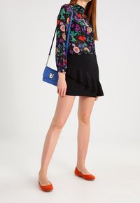 Schwarze Bluse mit langen Ärmeln und bunten Mustern, kombiniert mit einem schwarzen Rock mit Rüschensaum und orangefarbenen Ballerinas. Blauer Umhängetasche.