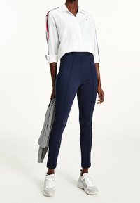 Valkoinen nappipaitapusero, jonka hihat on käärityt, tummansiniset legginssit ja harmaat lenkkarit. Legginseissä on sivusauman yksityiskohtia, ja paidassa on punaisia raitoja.