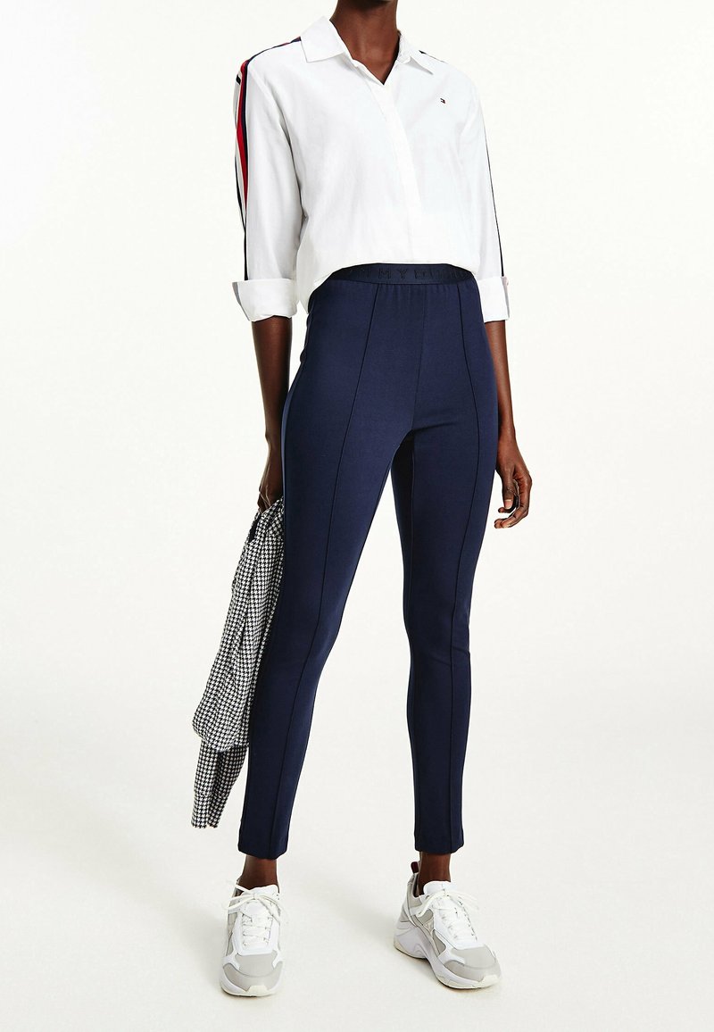 Valkoinen nappipaitapusero, jonka hihat on käärityt, tummansiniset legginssit ja harmaat lenkkarit. Legginseissä on sivusauman yksityiskohtia, ja paidassa on punaisia raitoja.