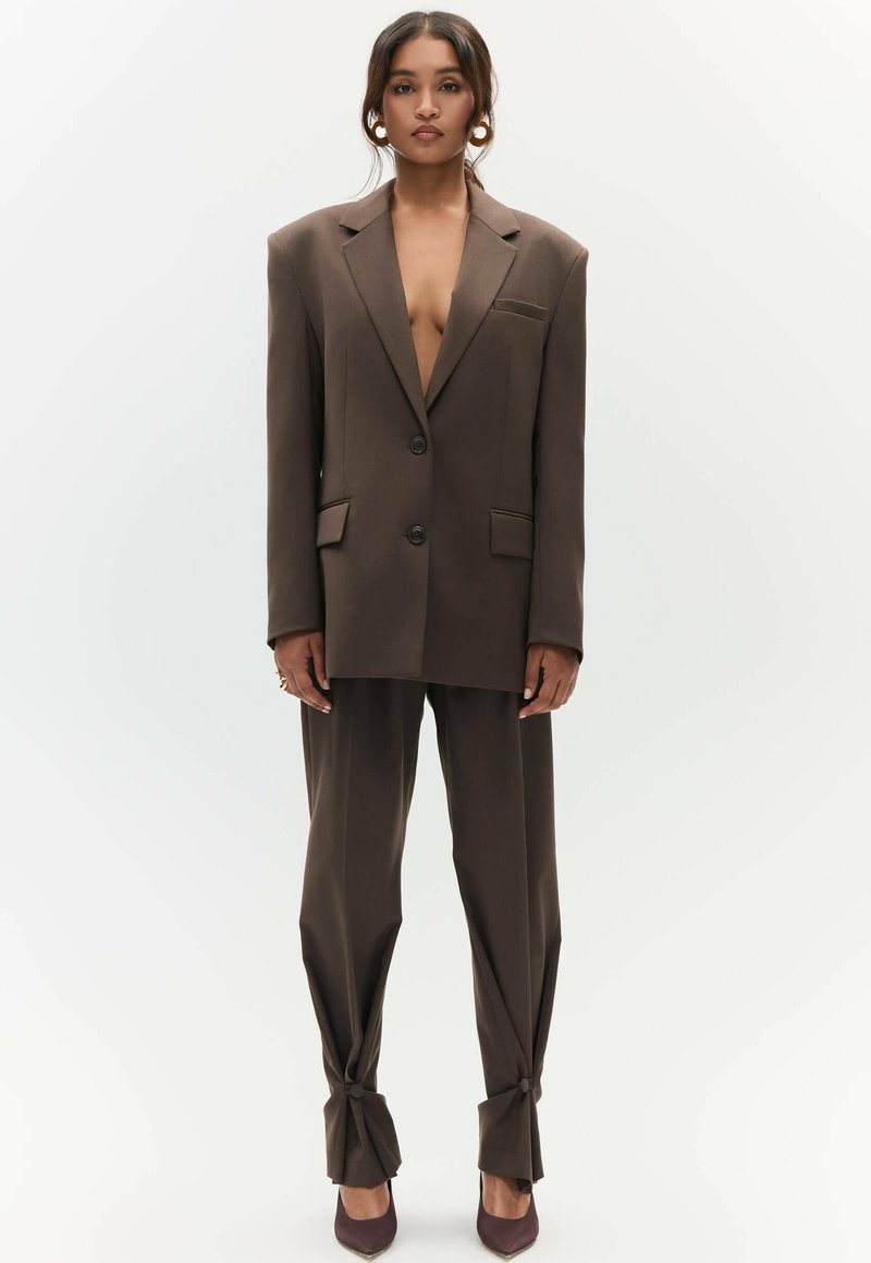 Bruin op maat gemaakt pak met een dubbelzijdige blazer met grote revers en broek met geknoopte manchet. Het materiaal lijkt glad en gestructureerd.