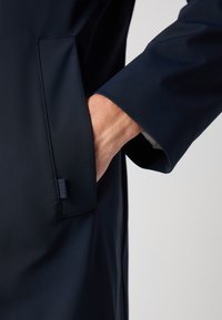 Cappotto impermeabile blu navy con una mano nella tasca laterale, design strutturato, texture liscia e un'etichetta sul bordo della tasca.