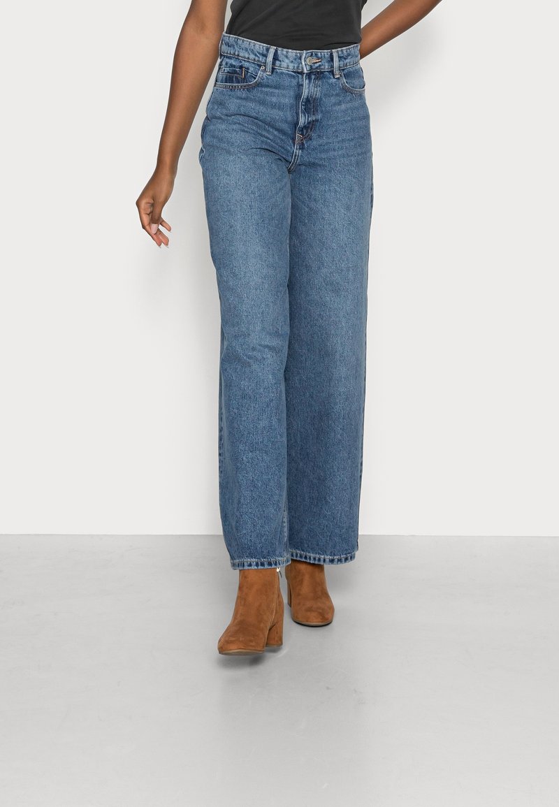 Jeans a gamba larga in denim blu con vita alta, caratterizzati da un design a cinque tasche e dettagli cuciti. Abbinati a stivali alla caviglia marroni.