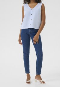 Top bianco senza maniche con strisce blu e bottoni dorati, abbinato a jeans blu attillati e sandali bianchi con tacco. Tessuto dalla consistenza liscia.
