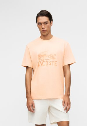 Joven con cabello oscuro vistiendo una camiseta Lacoste color melocotón con el logo del cocodrilo y pantalones cortos blancos con textura, posando frente a un fondo liso.